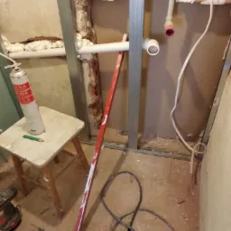 Montaż stelaża z profili stalowych do płyty karton gips, widoczne rury instalacyjne, piana montażowa, poziomica i wiertarka udarowa na podłodze.