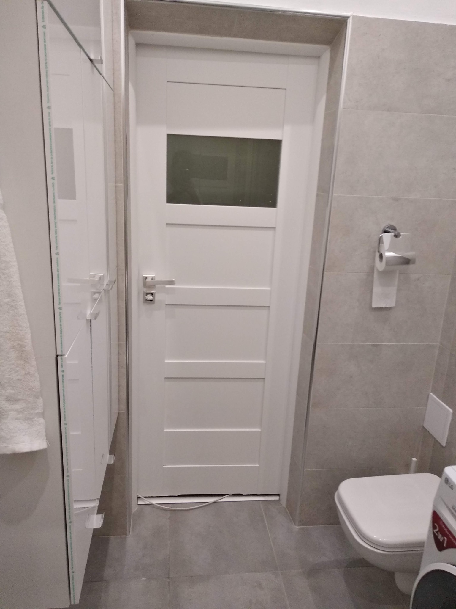 Biała, nowoczesna łazienka z drzwiami wewnętrznymi z szybą, szarą glazurą i podwieszaną toaletą.