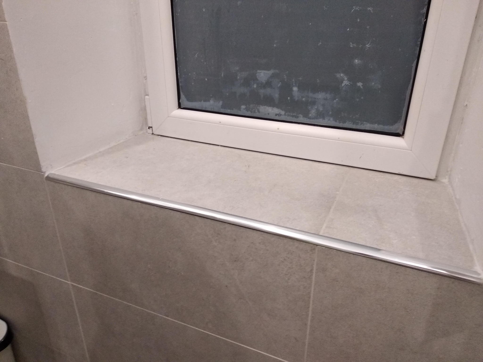 Szare płytki ceramiczne ułożone wokół okna z aluminiową listwą wykończeniową, widoczne fugi i zabrudzone okno.