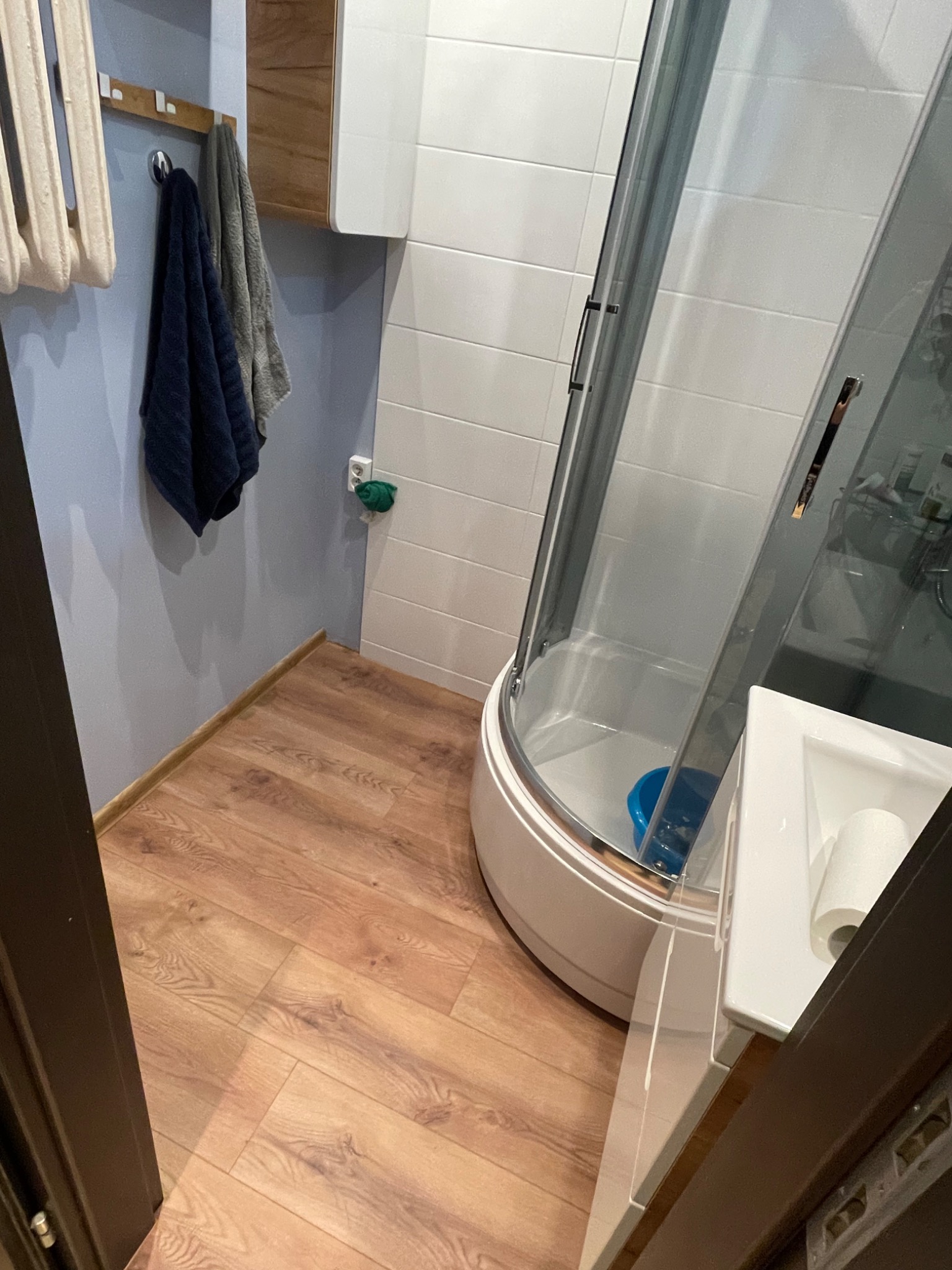 Widok na wykończoną łazienkę z kabiną prysznicową z zaokrąglonymi szybami, umywalką z rolką papieru toaletowego i drewnopodobną podłogą, widoczne ręczniki na wieszaku i grzejnik.