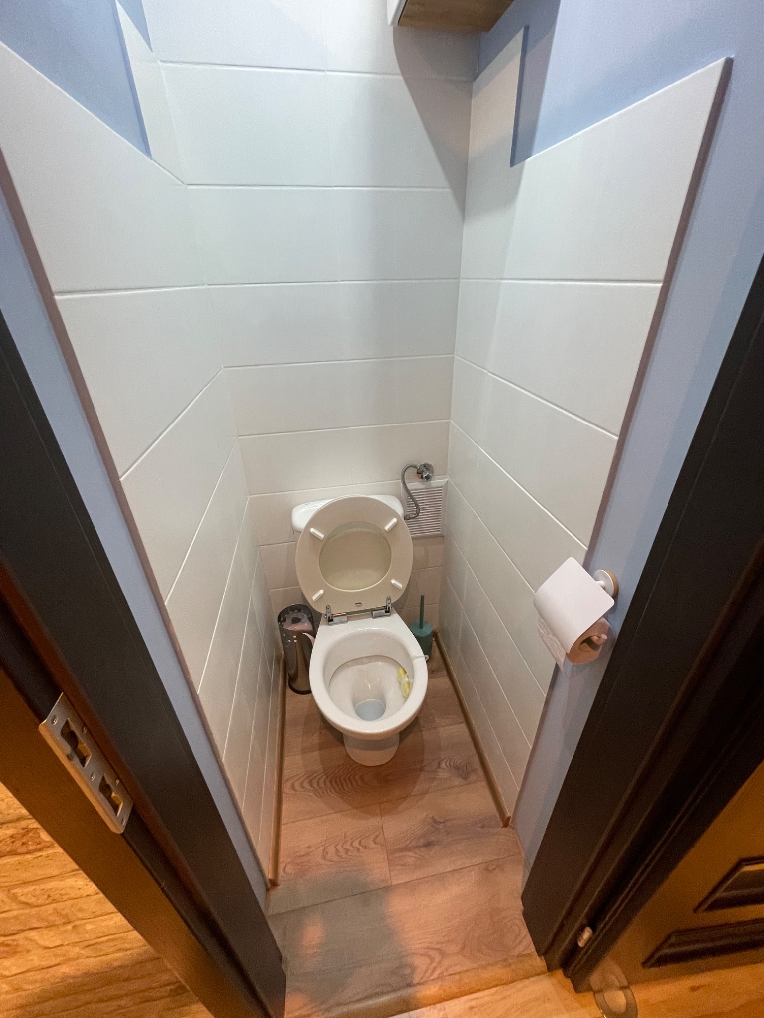 Wąska toaleta z drewnianą podłogą i białymi panelami ściennymi, widok zza otwartych, ciemnych drzwi.