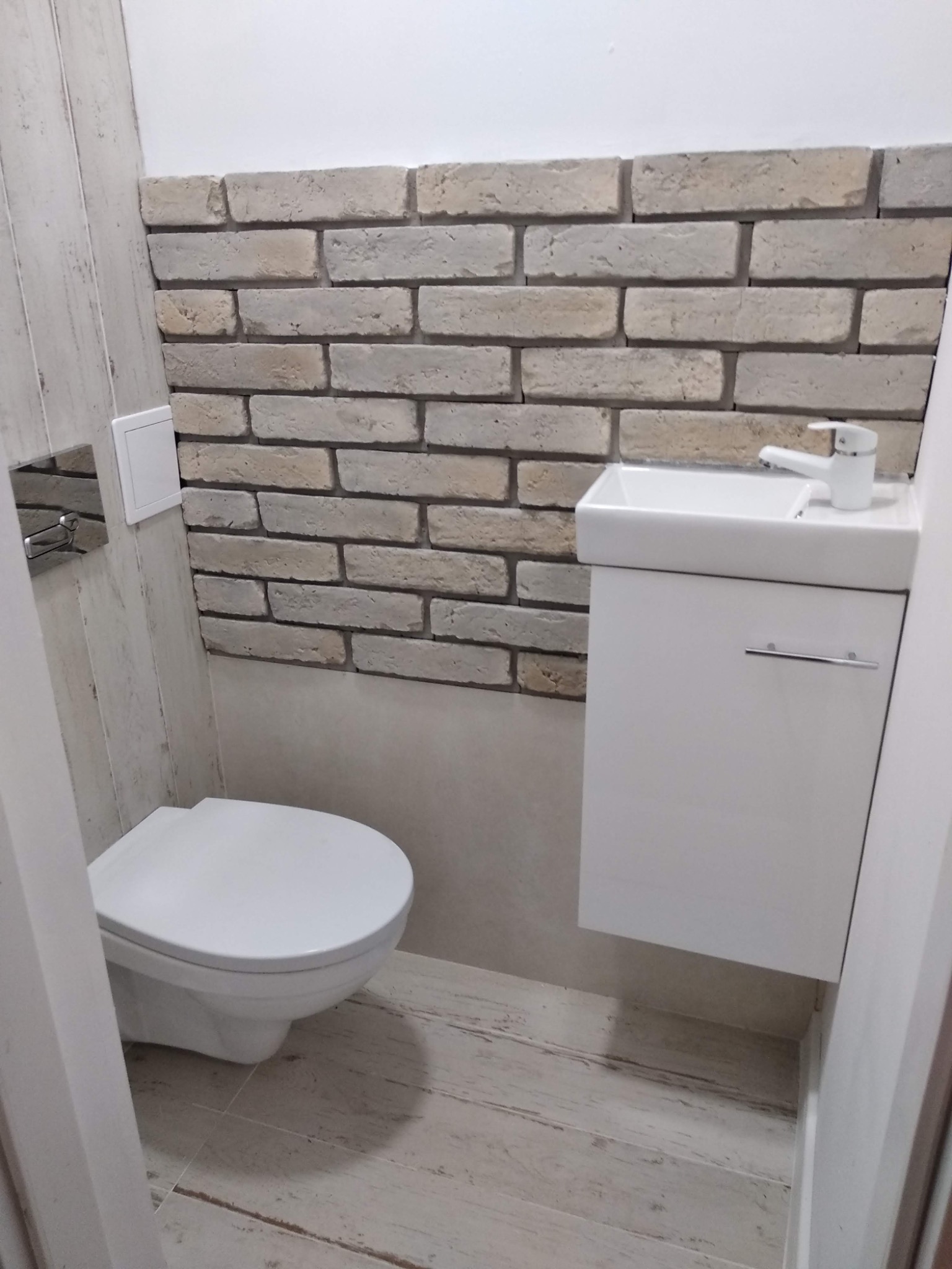 Mała łazienka z toaletą podwieszaną, umywalką nablatową na szafce i ścianą wykończoną dekoracyjną cegłą w odcieniach beżu i szarości.
