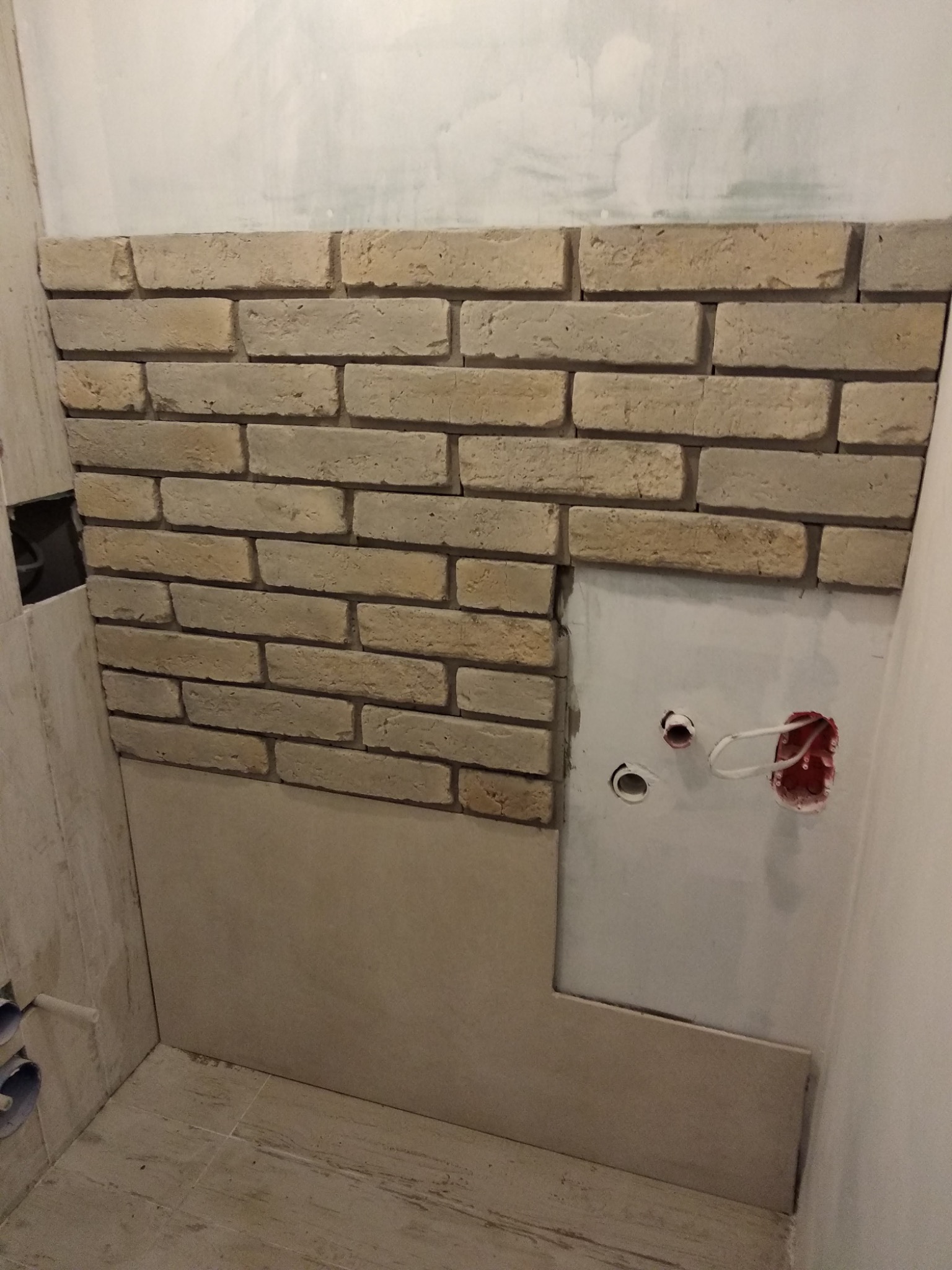 Fragment ściany łazienki w trakcie układania imitacji cegły, widoczne niedokończone fragmenty, odsłonięte instalacje elektryczne i hydrauliczne, oraz płytki podłogowe