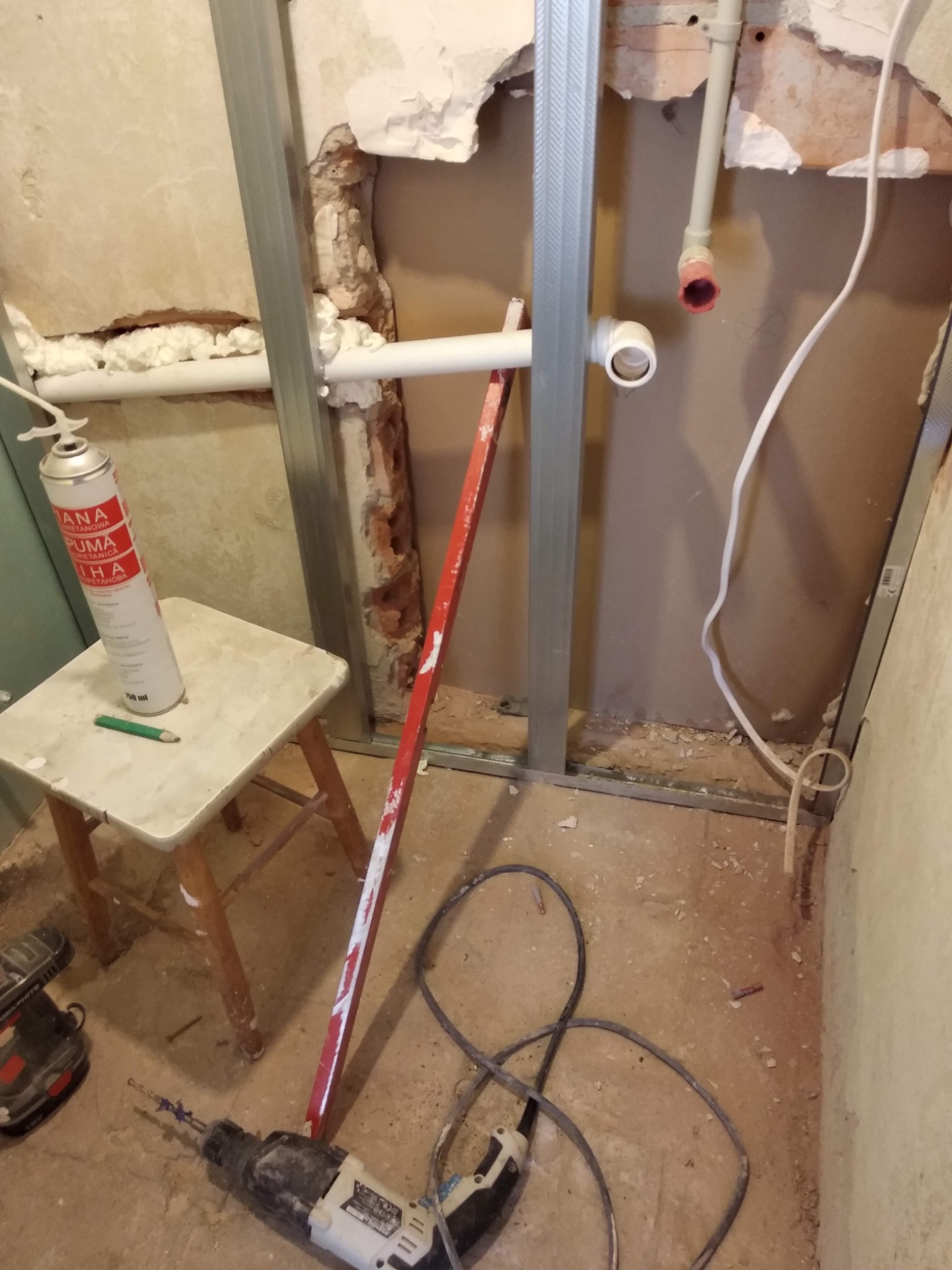 Montaż stelaża z profili stalowych do płyty karton gips, widoczne rury instalacyjne, piana montażowa, poziomica i wiertarka udarowa na podłodze.