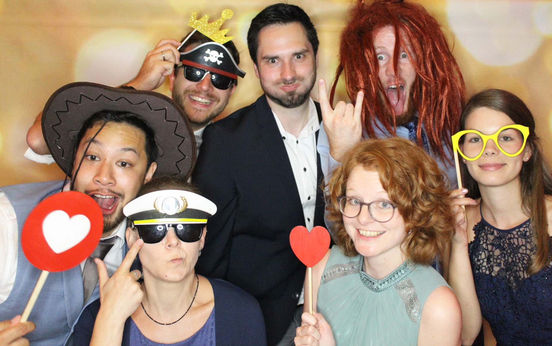 Grupa osób pozuje z rekwizytami: korona, piracka opaska, kowbojski kapelusz, czapka kapitana i serduszka na patyku, w tle jasne tło.