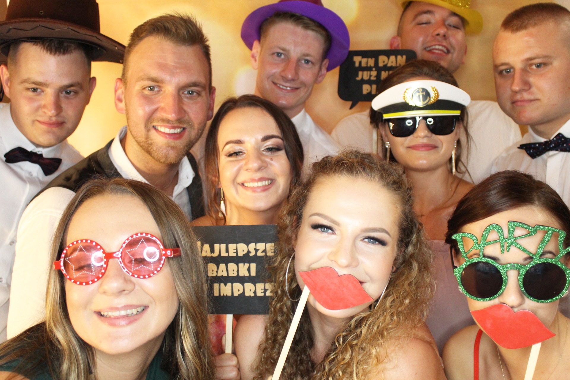 Grupa uśmiechniętych osób pozuje w fotobudce z rekwizytami: kapelusze, okulary z napisem 'Party', sztuczne usta na patyku, tabliczka z napisem 'Najlepsze babki imprezy'.