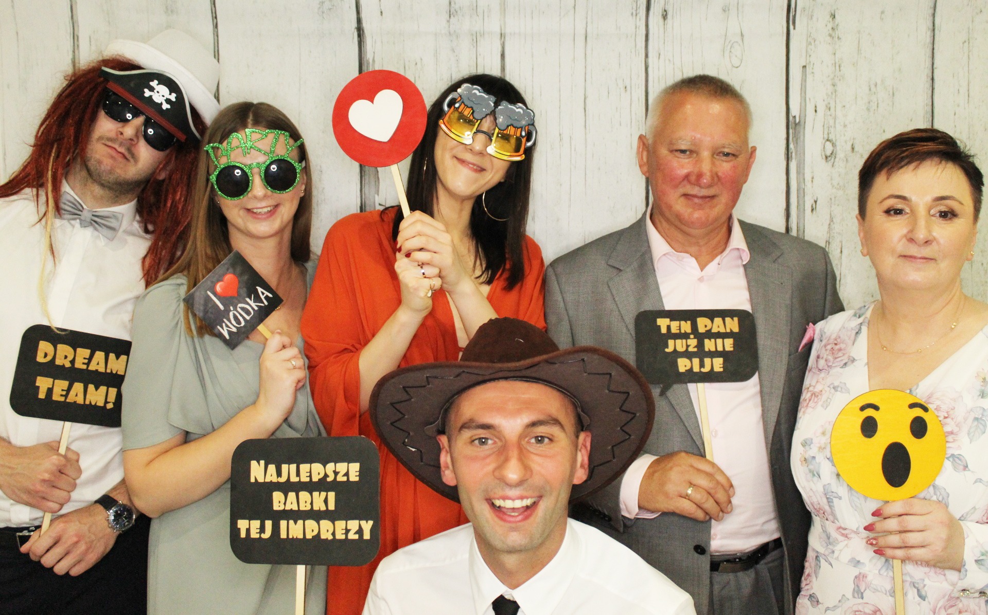 Grupa uśmiechniętych osób w przebraniach i z rekwizytami pozuje do zdjęcia na tle drewnianej ścianki.