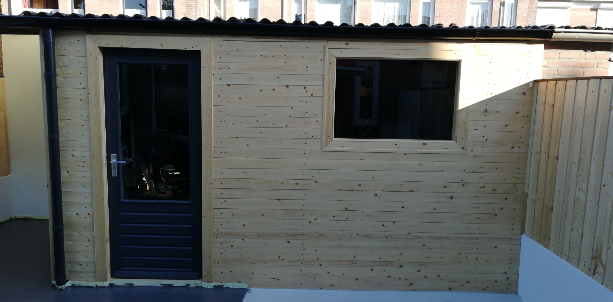 Drewniana konstrukcja szopy z zamontowanymi drzwiami w kolorze ciemnego grafitu i oknem, widoczne ślady po malowaniu na podłodze i ścianie, surowe drewno, bez wykończenia.