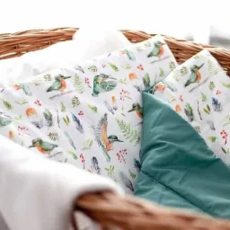 Wzór tkaniny z motywem akwarelowych ptaków i piór, umieszczony w wiklinowym koszu, obok gładkiej, turkusowej poduszki i białego kocyka.