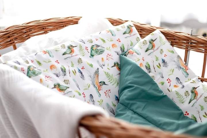 Wzór tkaniny z motywem akwarelowych ptaków i piór, umieszczony w wiklinowym koszu, obok gładkiej, turkusowej poduszki i białego kocyka.