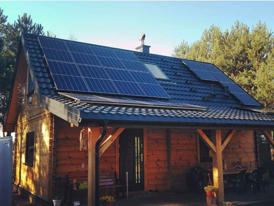 Drewniany dom z ciemną, metalową dachówką, na której zamontowano panele słoneczne, z widocznym kominem zwieńczonym ozdobną figurką, na tle zielonych drzew.