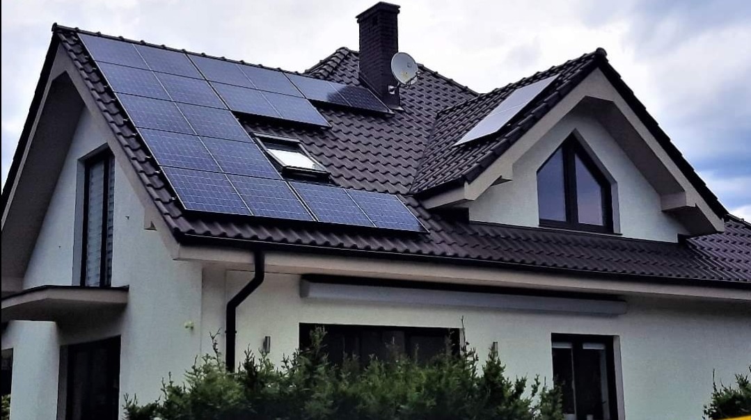 Panele słoneczne na dachu dwuspadowym z dachówką ceramiczną, widoczne okno dachowe i antena satelitarna.