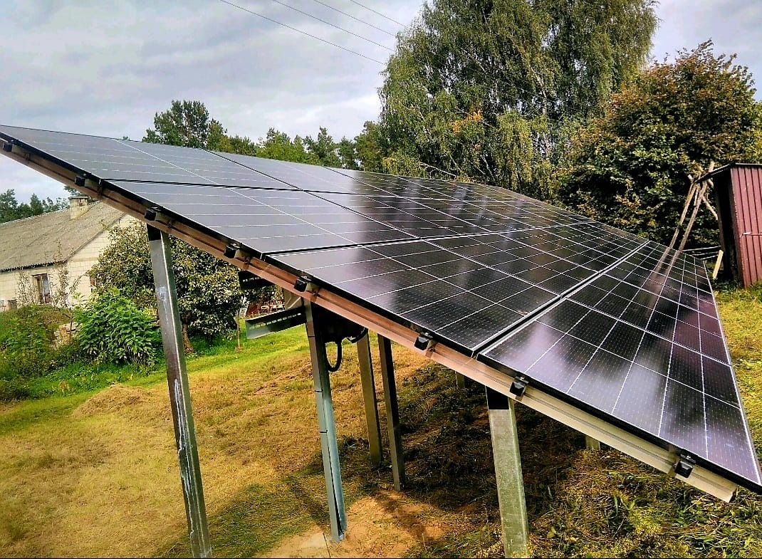 Panel słoneczny zamontowany na metalowej konstrukcji, z widocznym domem i drzewami w tle, na lekko pochylonym terenie.