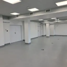 Wnętrze nowo wybudowanej hali lub biura z szarą podłogą, białymi ścianami, kolumnami i drzwiami, widoczne kratki wentylacyjne pod sufitem i klimatyzatory.