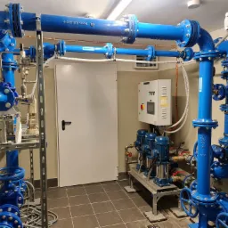 Węzeł hydroforowy z czterema pompami pionowymi w niebieskiej obudowie, rozbudowany system rur z licznymi zaworami, manometrami i osprzętem, biała szafa sterownicza na ścianie, białe drzwi w tle.