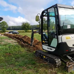 Mini koparka Schaeff SV18 w trakcie wykonywania wykopu liniowego na trawiastym terenie, widoczne wykopane bloczki i samochód w tle, błękitne niebo z chmurami.