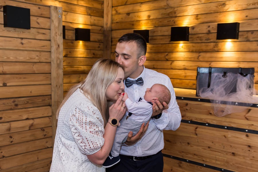 Rodzice z noworodkiem w eleganckich ubraniach pozują na tle drewnianej ściany z czarnymi lampami, mama trzyma rączkę dziecka przy ustach, tata całuje mamę w głowę.