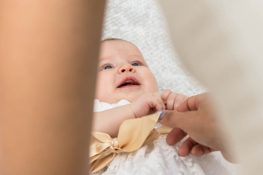 Uśmiechnięty noworodek z niebieskimi oczami, leżący na białym kocyku w białej sukience z kokardą, trzymany za rączkę przez osobę z fioletowym lakierem na paznokciach, uchwycony z bliska...