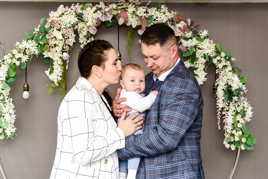 Rodzinne zdjęcie: matka całuje niemowlę trzymane przez ojca w eleganckim, kwiatowym łuku z wiszącymi żarówkami.