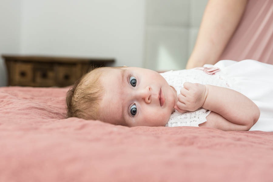 Niemowlę w białej, koronkowej sukience leży na różowym kocyku, z zaciśniętą piąstką przy ustach i szeroko otwartymi, niebieskimi oczami, spoglądając w obiektyw.