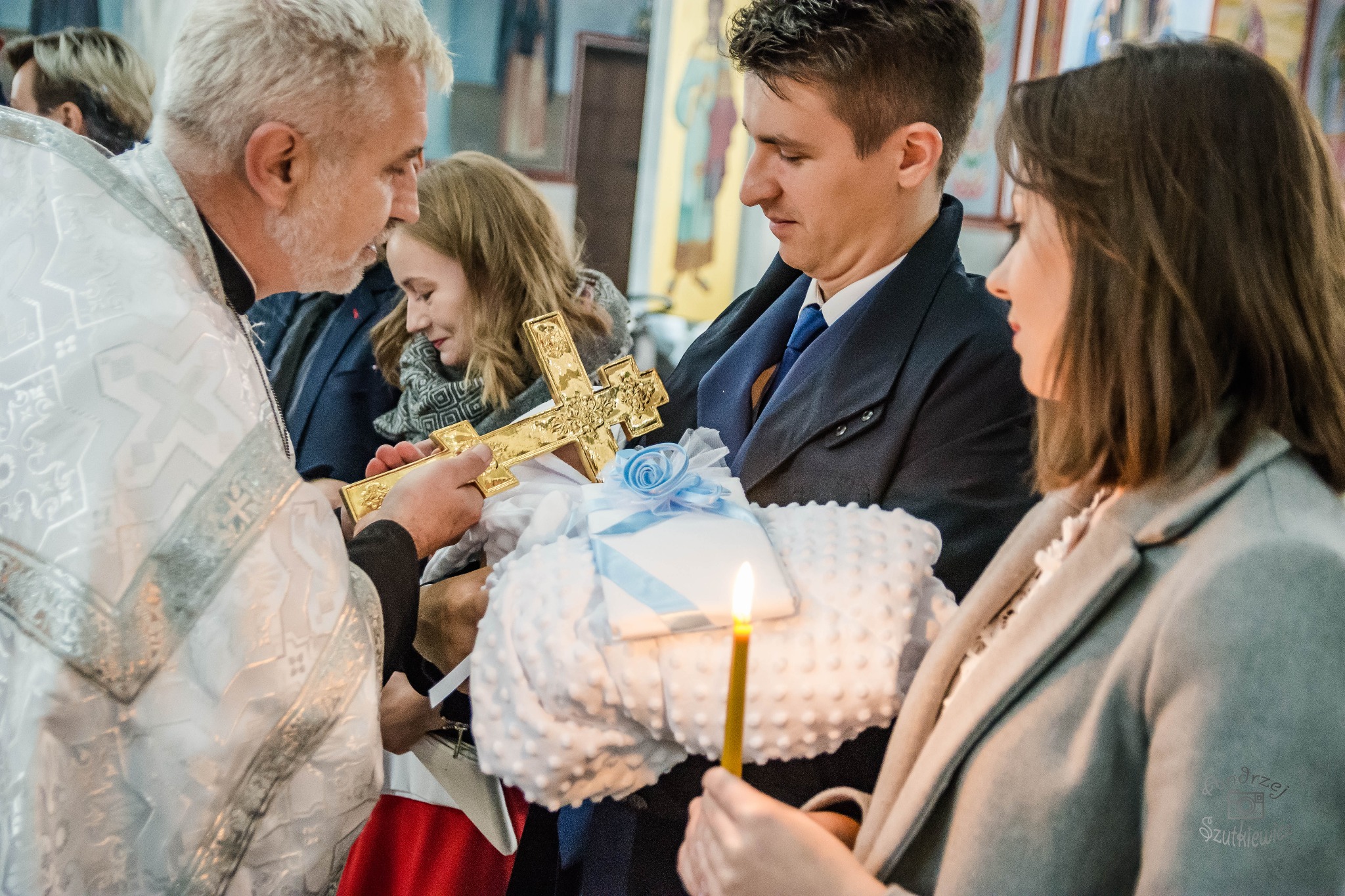 Ksiądz w bogato zdobionej szacie trzyma złoty krzyż nad dzieckiem w białym rożku z niebieską wstążką, otoczonym przez rodziców z zapaloną świecą w dłoni.