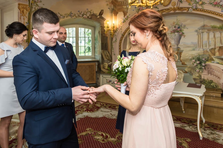 Moment zakładania obrączki podczas ceremonii ślubnej w bogato zdobionym wnętrzu, panna młoda w pudrowej sukni z koronkowym wykończeniem, pan młody w granatowym garniturze z muchą.