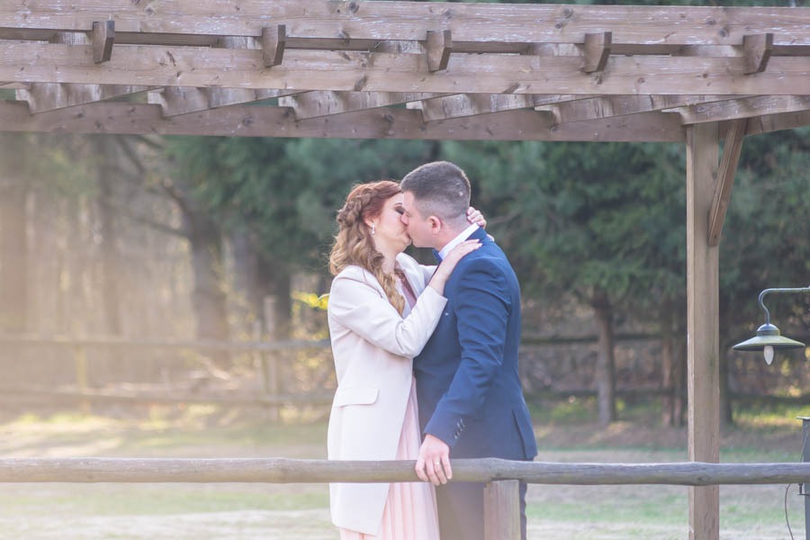 Para młoda całuje się pod drewnianą pergolą w plenerze, ona w jasnoróżowym płaszczu, on w granatowym garniturze, w tle drzewa i delikatne światło słoneczne.