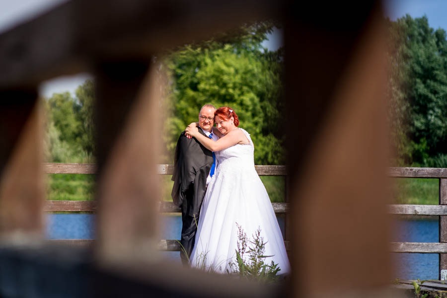 Para młoda w objęciach, widziana przez drewniane balustrady mostu, z zielonym tłem drzew i wody.