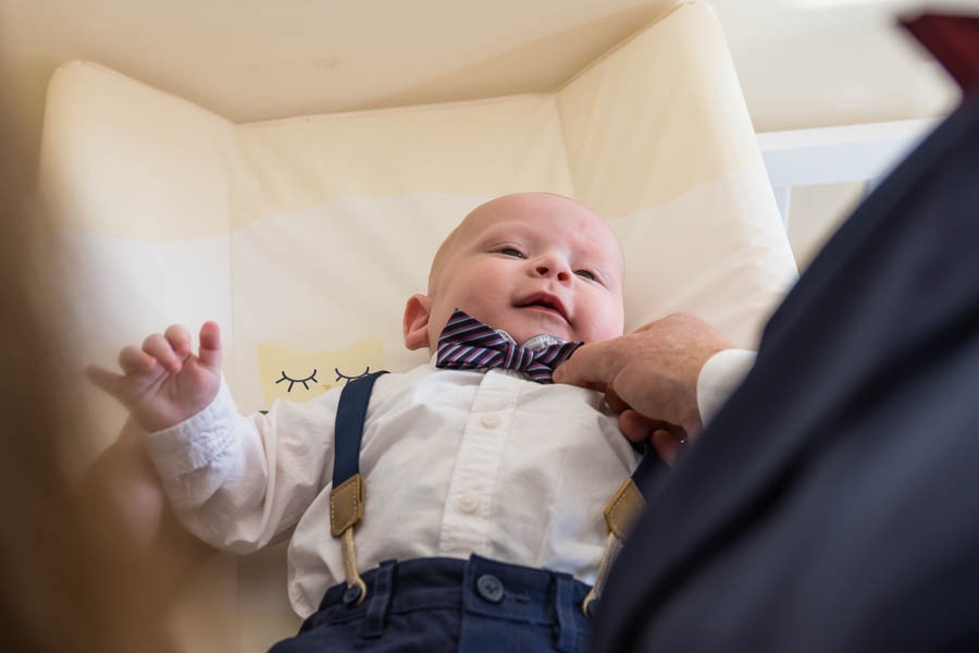 Uśmiechnięty niemowlak w koszuli, spodniach na szelkach i muszce, układanej przez dłoń w mankiecie marynarki, leży na przewijaku z motywem śpiącego kotka.