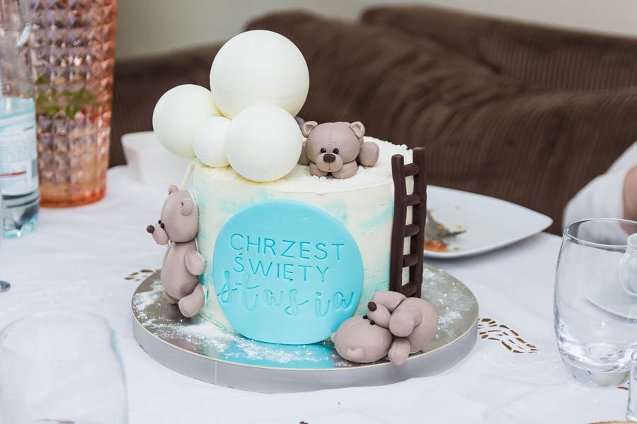 Tort na chrzest ozdobiony niebieskim napisem, białymi balonami, czekoladową drabinką i figurkami misiów z lukru.