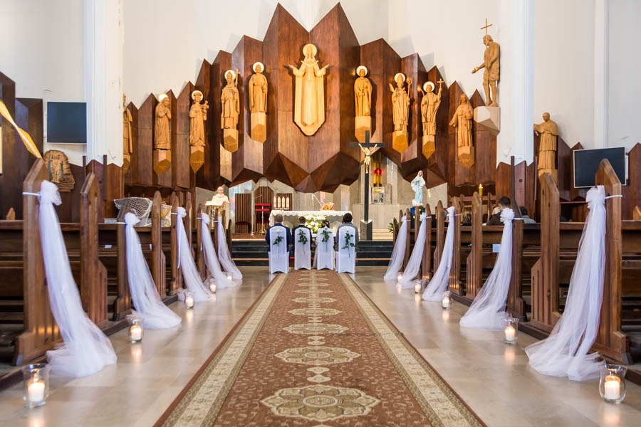 Wnętrze kościoła przygotowane na uroczystość, z dekoracjami z białego tiulu na ławkach i świecami w szklanych lampionach wzdłuż dywanu prowadzącego do ołtarza, gdzie widać sylwetki osób w jasnych...