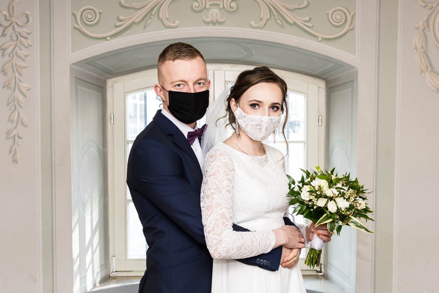 Para młoda w maseczkach ochronnych pozuje do zdjęcia ślubnego w jasnym wnętrzu, panna młoda trzyma bukiet białych kwiatów.