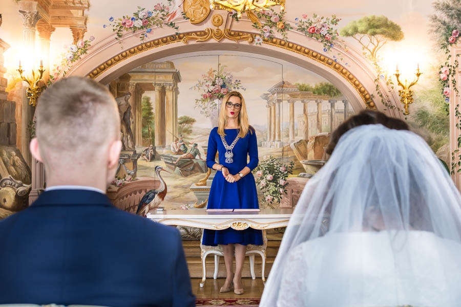 Urzędniczka w niebieskiej sukience i łańcuchu w sali ślubów z freskami, podczas ceremonii zaślubin pary młodej.