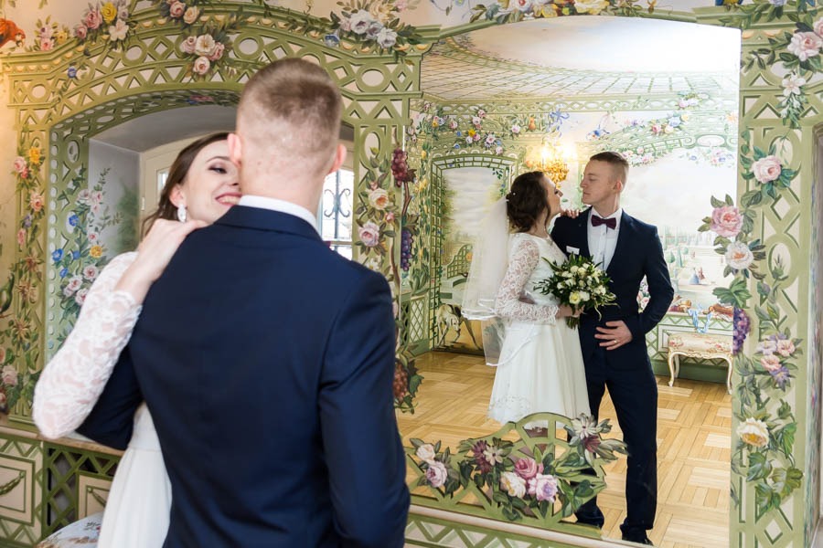 Para młoda w dekoracyjnym wnętrzu z motywami kwiatowymi, panna młoda w koronkowej sukni obejmuje pana młodego, który stoi tyłem do obiektywu, w tle odbicie pary w podobnej pozie.
