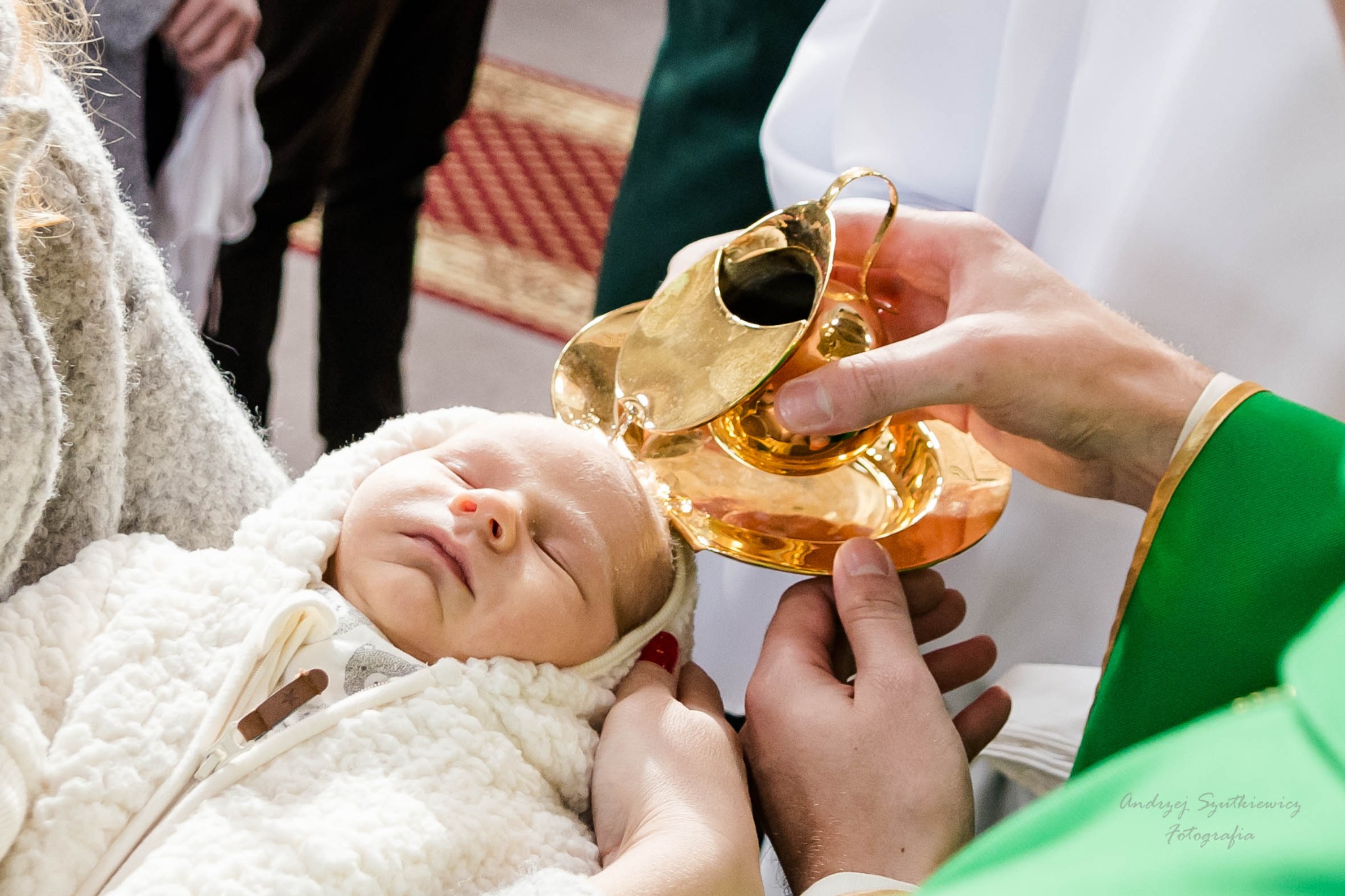 Ksiądz polewa wodą święconą śpiące niemowlę ubrane w biały kombinezon z kapturem, trzymane przez kobietę z czerwonym lakierem na paznokciach.