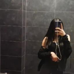 Osoba robi selfie w łazience z szarymi płytkami, widoczna część sylwetki w czarnym ubraniu, słuchawki w uszach.