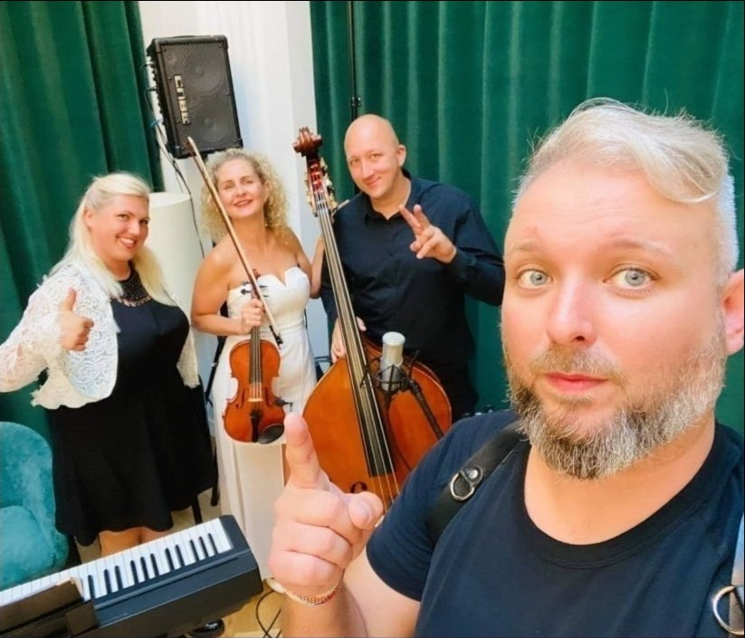 Czteroosobowy zespół muzyczny w trakcie selfie: kobieta z skrzypcami, mężczyzna z kontrabasem, kobieta dająca kciuk w górę i mężczyzna robiący zdjęcie, widoczne również fragmenty klawiszy...