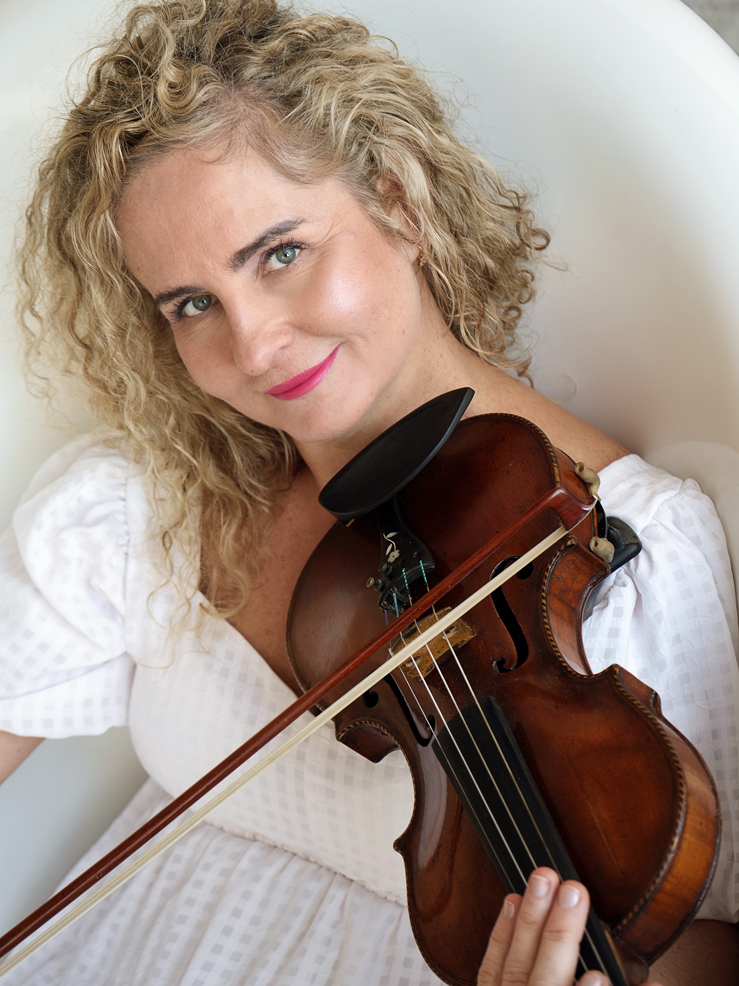 Portret kobiety z blond lokami w białej sukience, trzymającej skrzypce i smyczek, spoglądającej w obiektyw z lekkim uśmiechem.