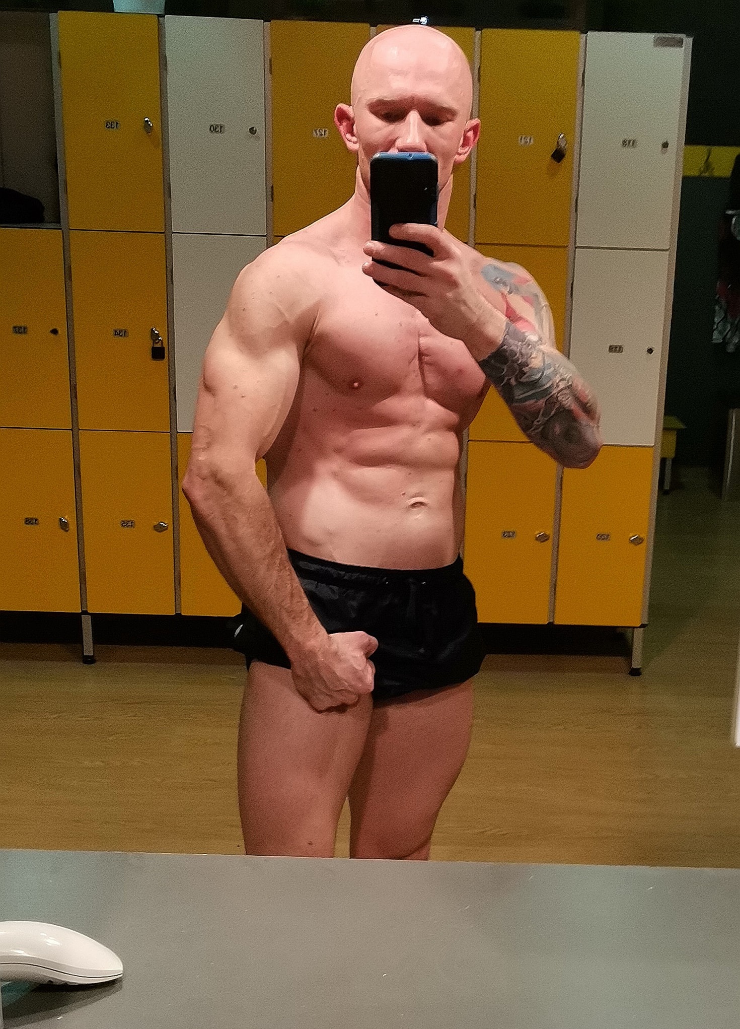 Mężczyzna bez włosów, z tatuażem na ramieniu, robi selfie w szatni na tle żółto-białych szafek.