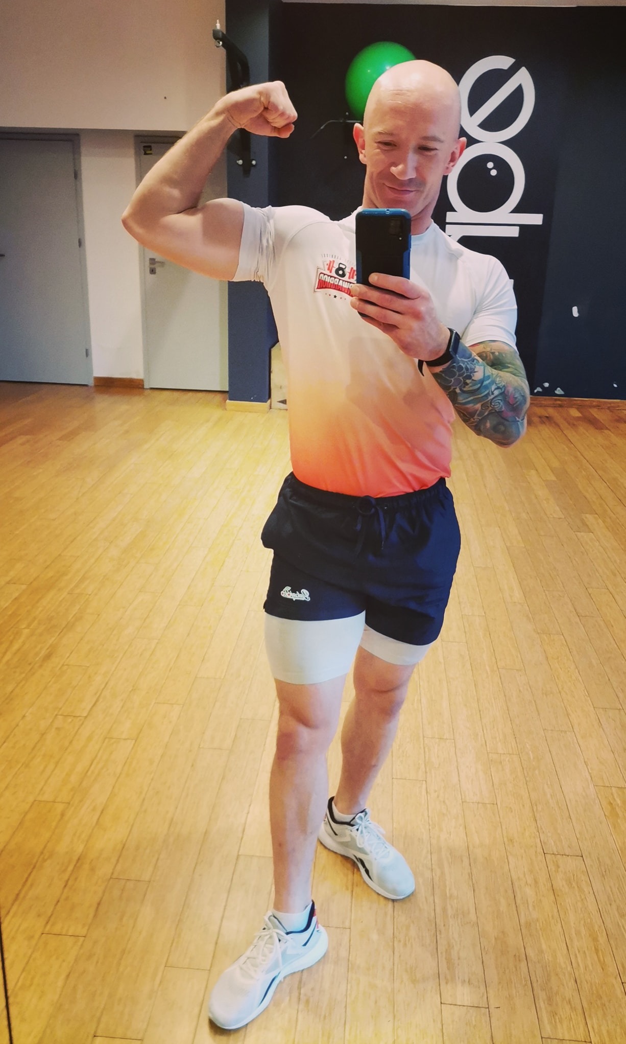 Mężczyzna w sportowym stroju robi selfie, prezentując biceps na tle sali gimnastycznej z widocznym logo na ścianie i zieloną piłką do ćwiczeń.