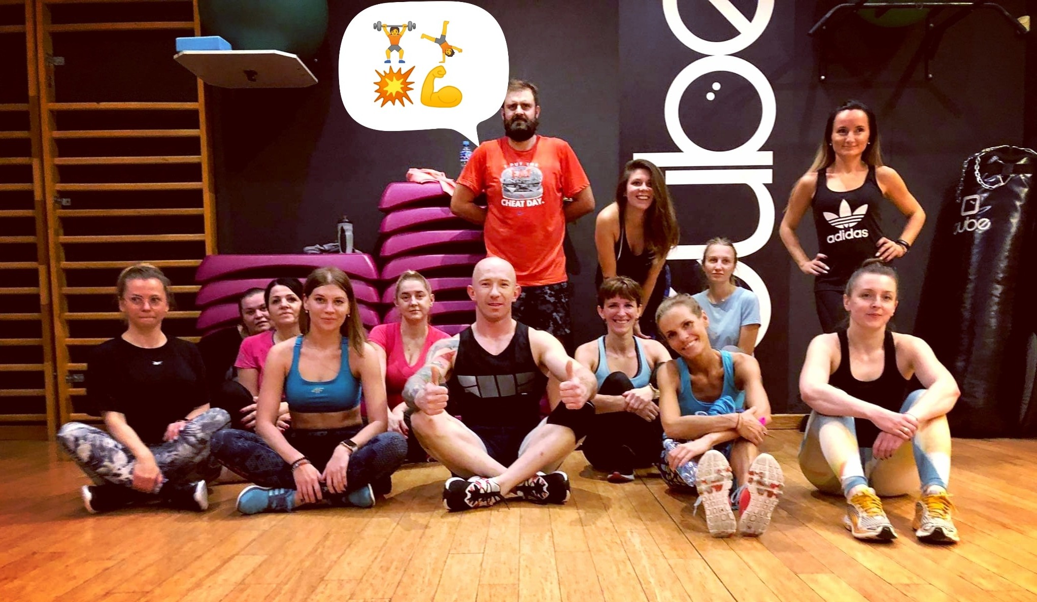 Grupa osób siedzi na podłodze w sali gimnastycznej, z mężczyzną w środku pokazującym kciuki w górę, w tle drabinka gimnastyczna i worki treningowe.