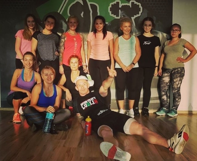 Grupa kobiet w strojach sportowych pozuje z mężczyzną w koszulce 'Sportowy Lublin' leżącym na podłodze po treningu w siłowni.