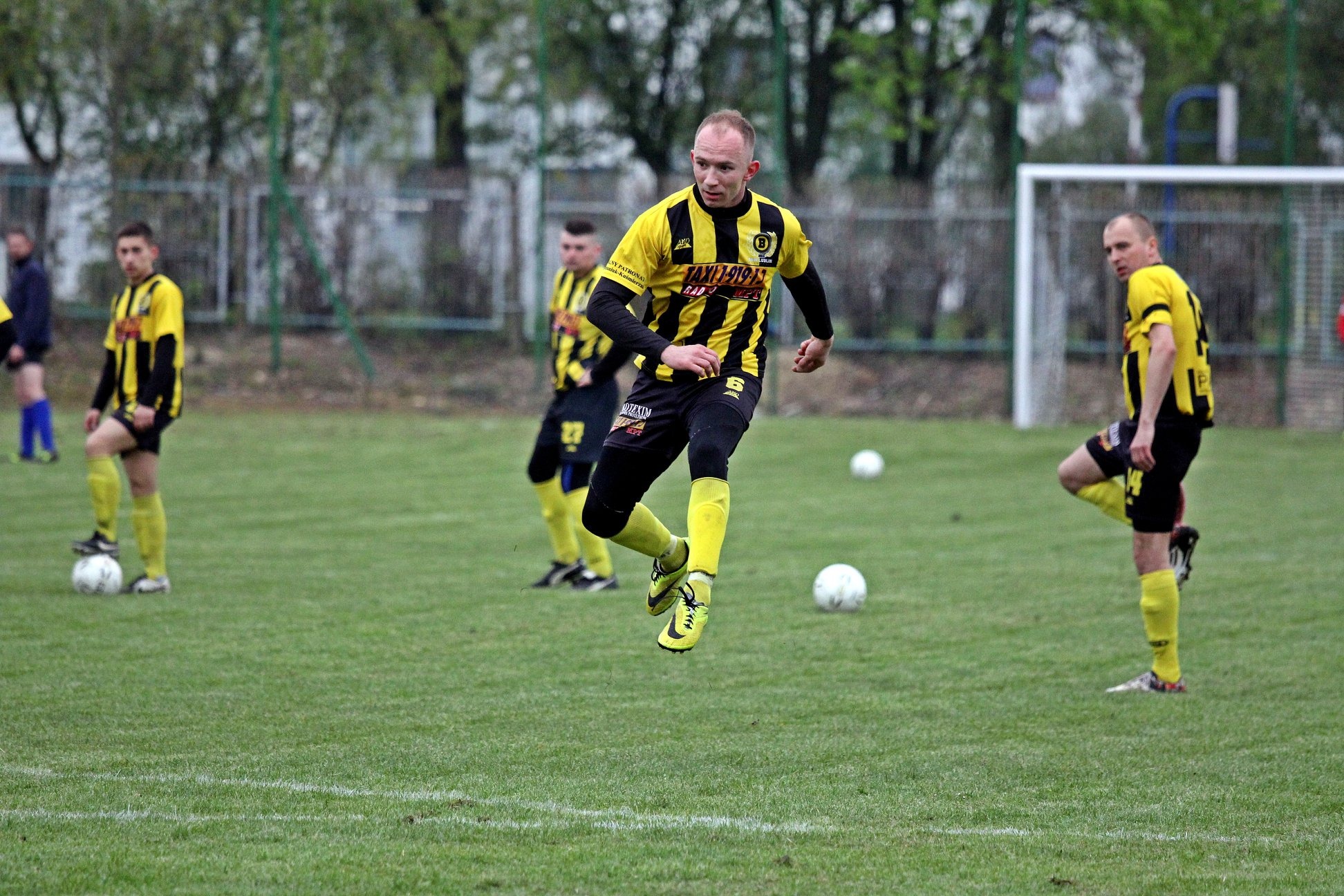 Piłkarz w żółto-czarnym stroju w trakcie skoku na boisku podczas meczu, w tle inni zawodnicy i bramka.