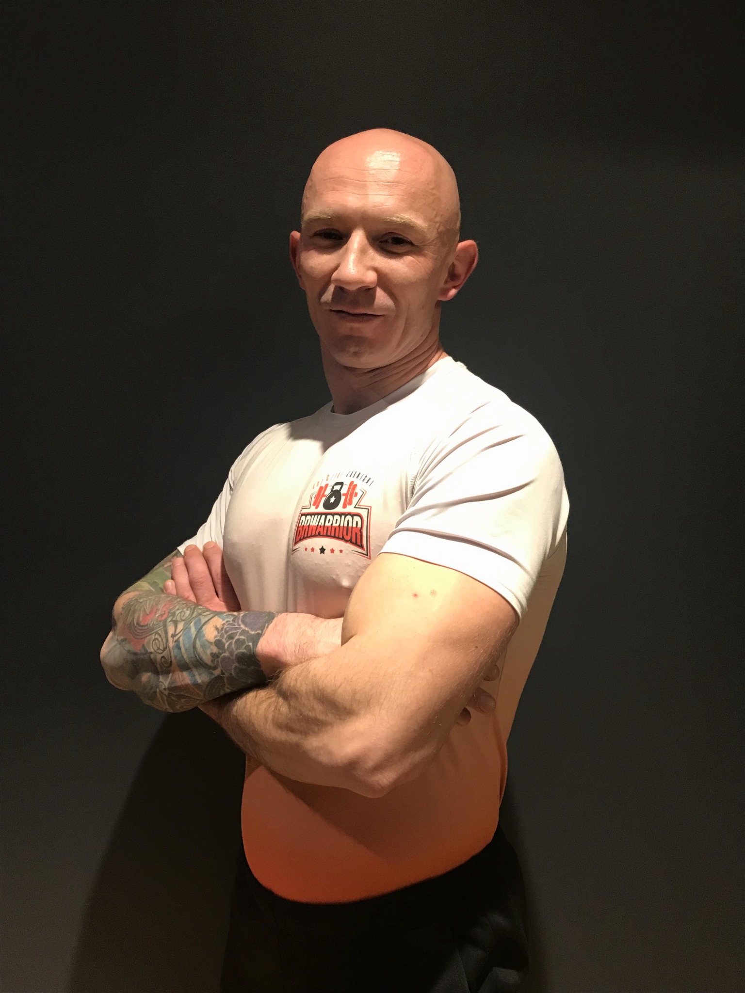 Uśmiechnięty, łysy mężczyzna z tatuażami na ramieniu, ubrany w białą koszulkę z logo 'Row Warrior', stoi na tle ciemnoszarej ściany z założonymi rękami.