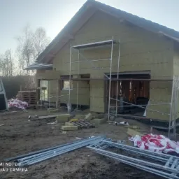 Dom w trakcie ocieplania, widoczne rusztowania, materiały izolacyjne, częściowo ocieplona elewacja, teren budowy.