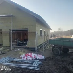 Budowa domu jednorodzinnego w trakcie, widoczne rusztowania, przyczepa rolnicza i ocieplone ściany zewnętrzne płytami izolacyjnymi.