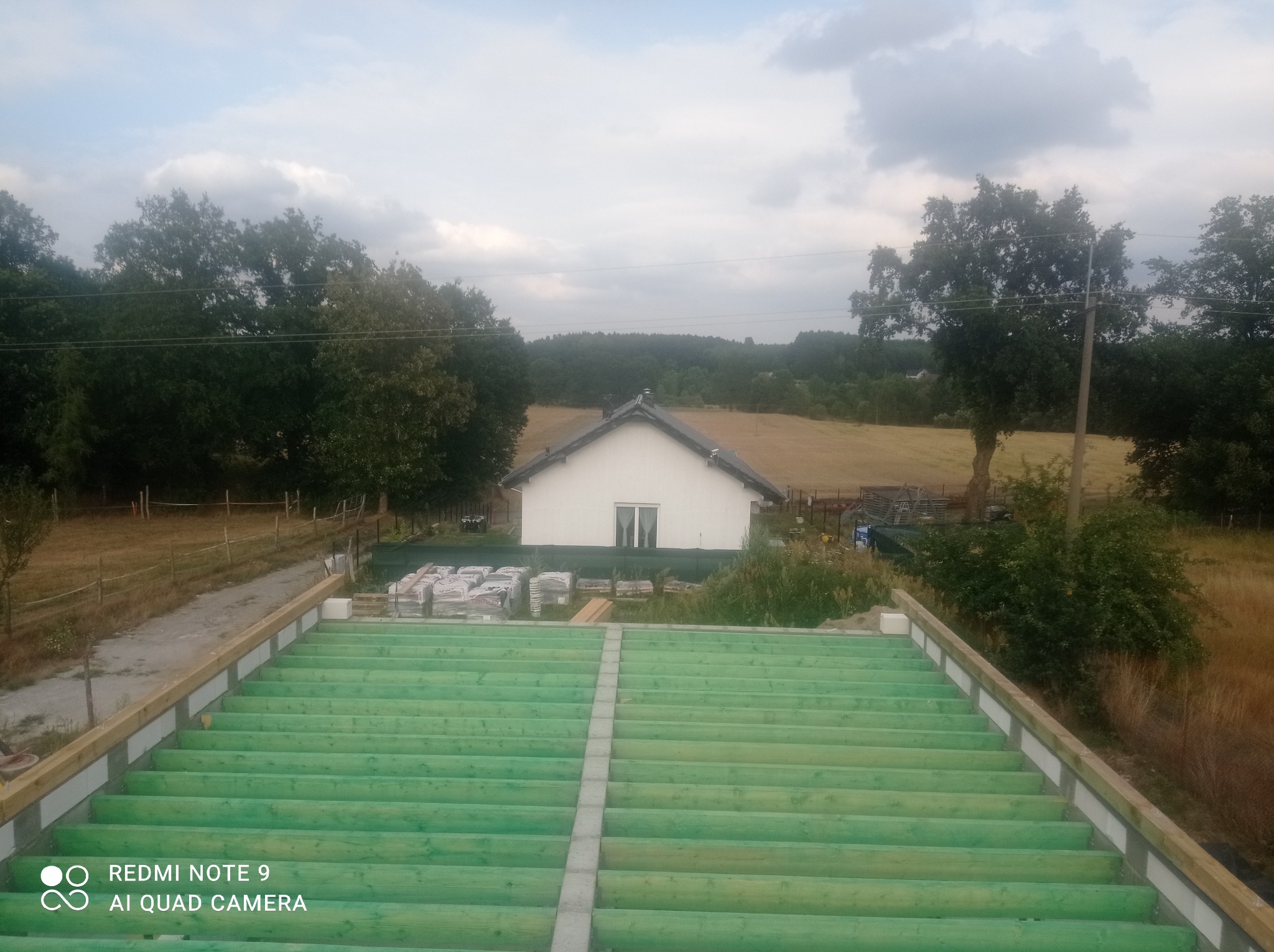 Konstrukcja stropu z zielonych belek drewnianych na tle budowanego domu i krajobrazu polno-leśnego, widok z góry.