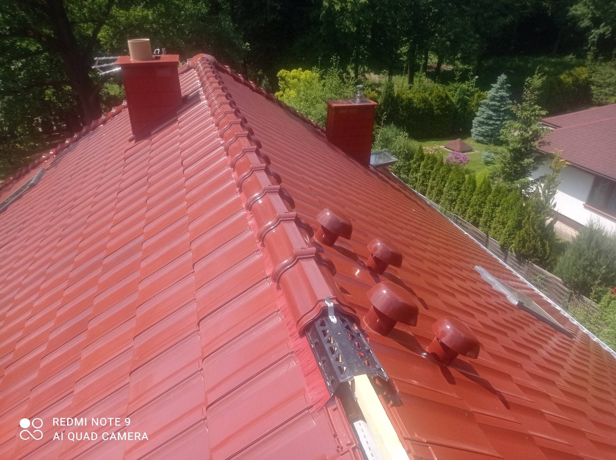 Czerwony dach z dachówki ceramicznej, widoczne kominy i elementy wentylacyjne, w tle zieleń drzew i krzewów.