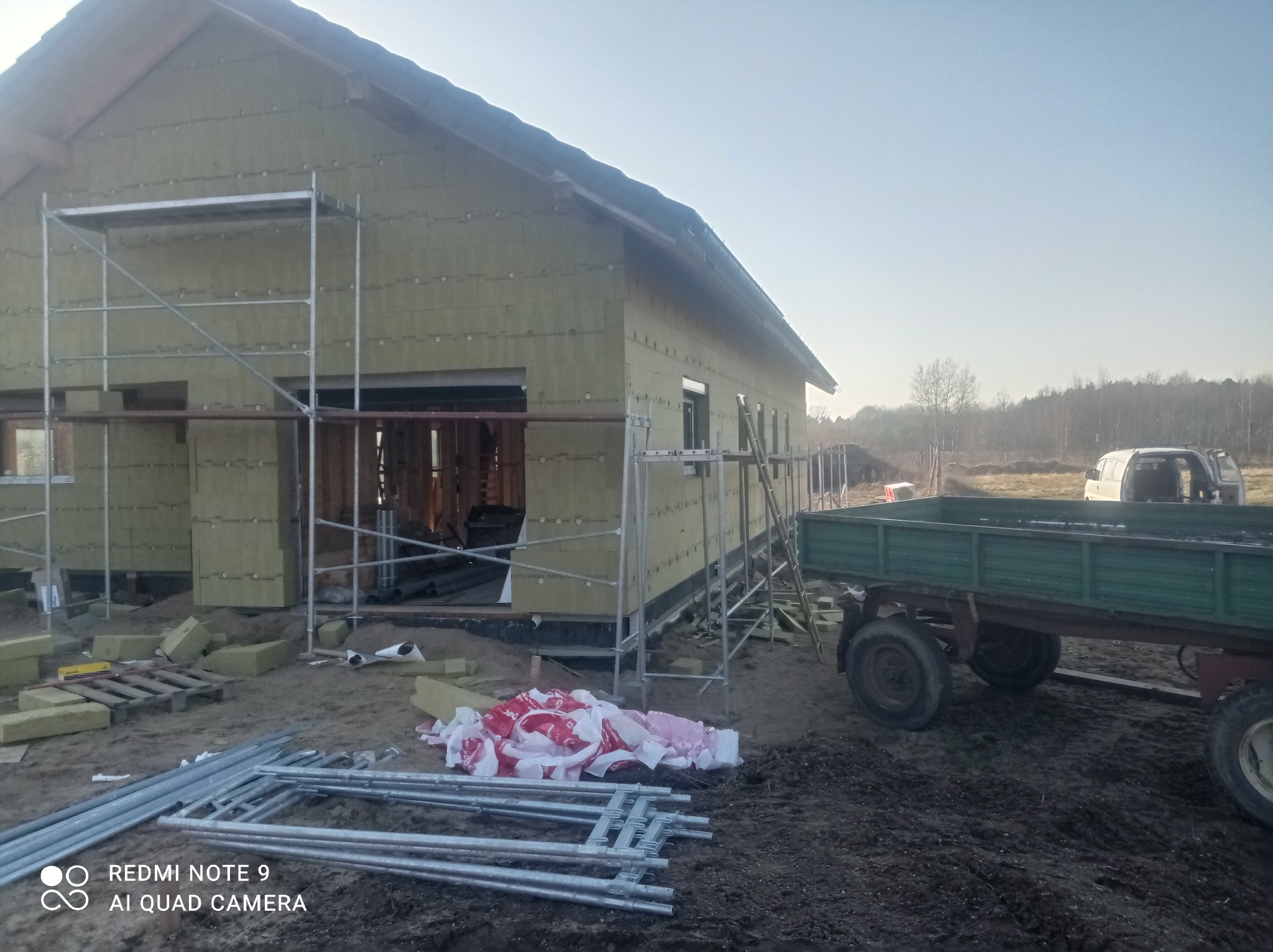 Budowa domu jednorodzinnego w trakcie, widoczne rusztowania, przyczepa rolnicza i ocieplone ściany zewnętrzne płytami izolacyjnymi.