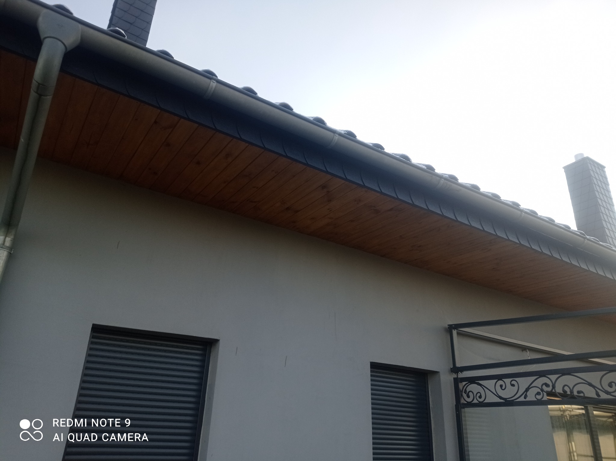 Nowoczesny dom z szarą elewacją, ciemnymi roletami okiennymi i drewnianą podbitką dachową, widoczny fragment dachu z orynnowaniem i komin w tle.